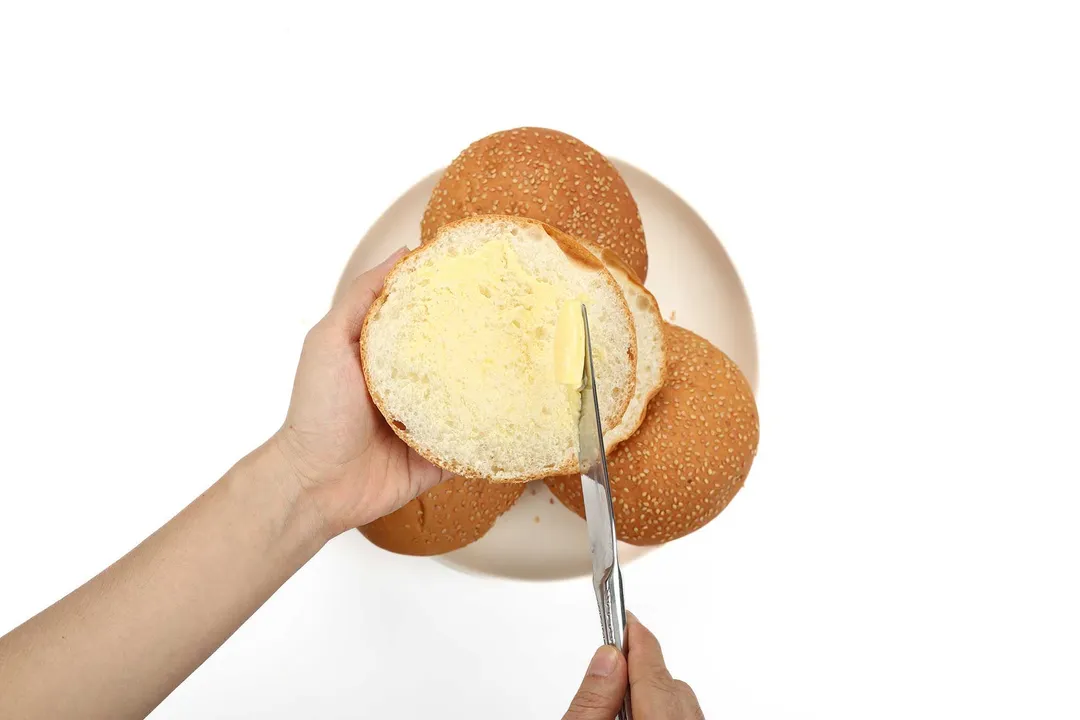 A hand holding up half a burger bun while the other hand sweeps butter onto the bun's inner surface.