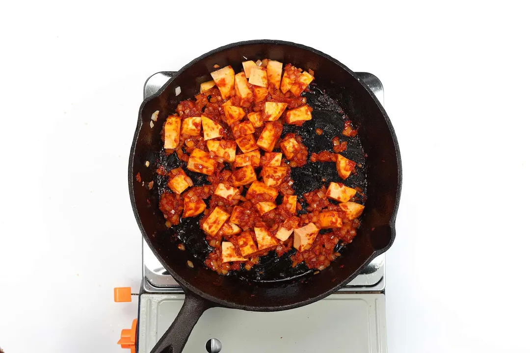 A skillet cooking sweet potato cubes covered in tomato paste on a portable gas stove.