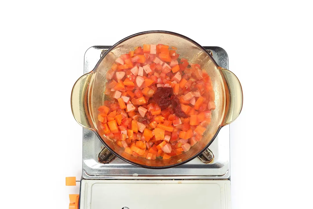 A saucepan cooking diced vegetables and sausage cubes with a dollop of tomato paste in the center.