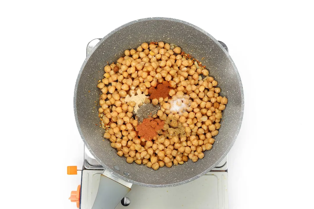 A pan cooking chickpea with various condimens on a portable gas stove.