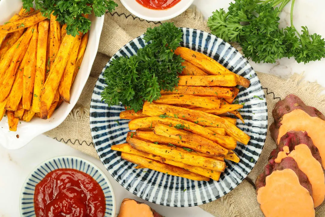 two plates of cooked sweet potato sticks decorated with ketchup bowls and slices sweet potato, parsley