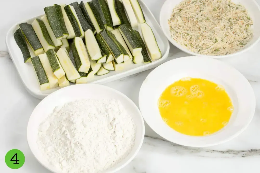 Plates of ingredients: zucchini fingers, beaten eggs, flour, and breadcrumb mixture