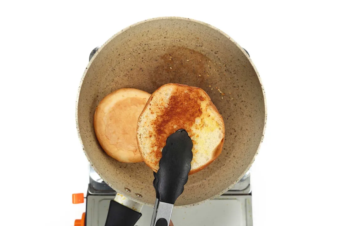 A pair of tongs picking up half a burger bun that's been cooked on a large pan.