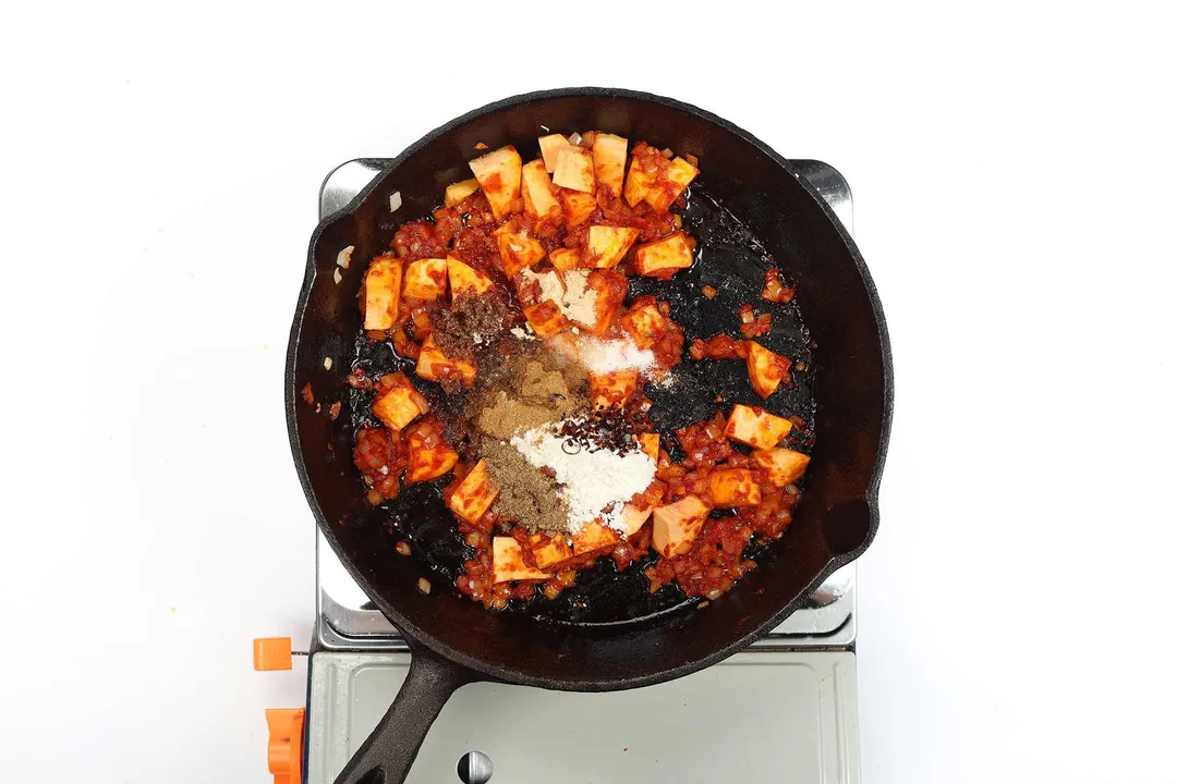 A skillet cooking sweet potato cubes covered in tomato paste with spices and seasonings.