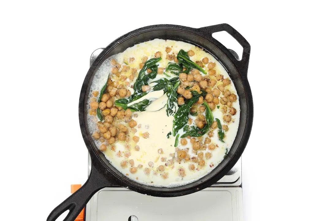 A skillet cooking chickpeas and spinach with heavy cream and milk on a portable gas stove