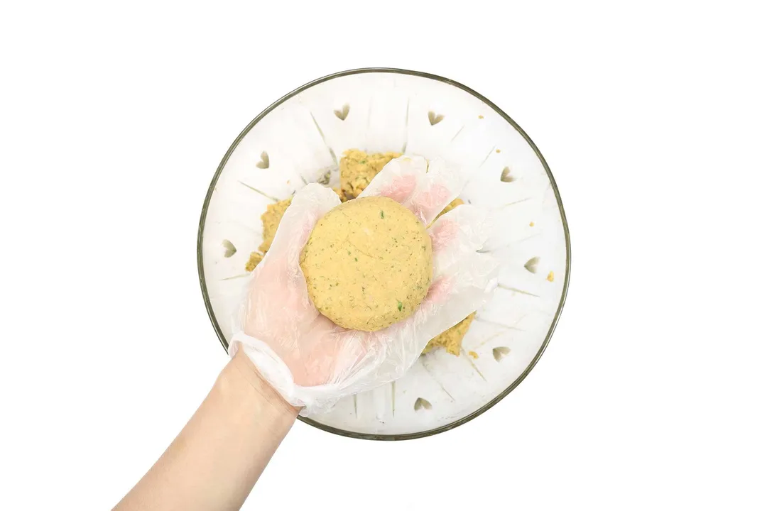 A hand wearing a disposable plastic glove holding a pattie of blended chickpeas.