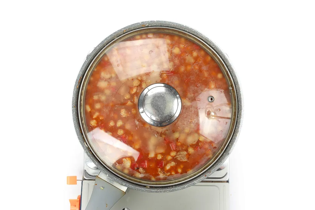A large pan cooking chickpea rice in tomato sauce covered with a glass lid.