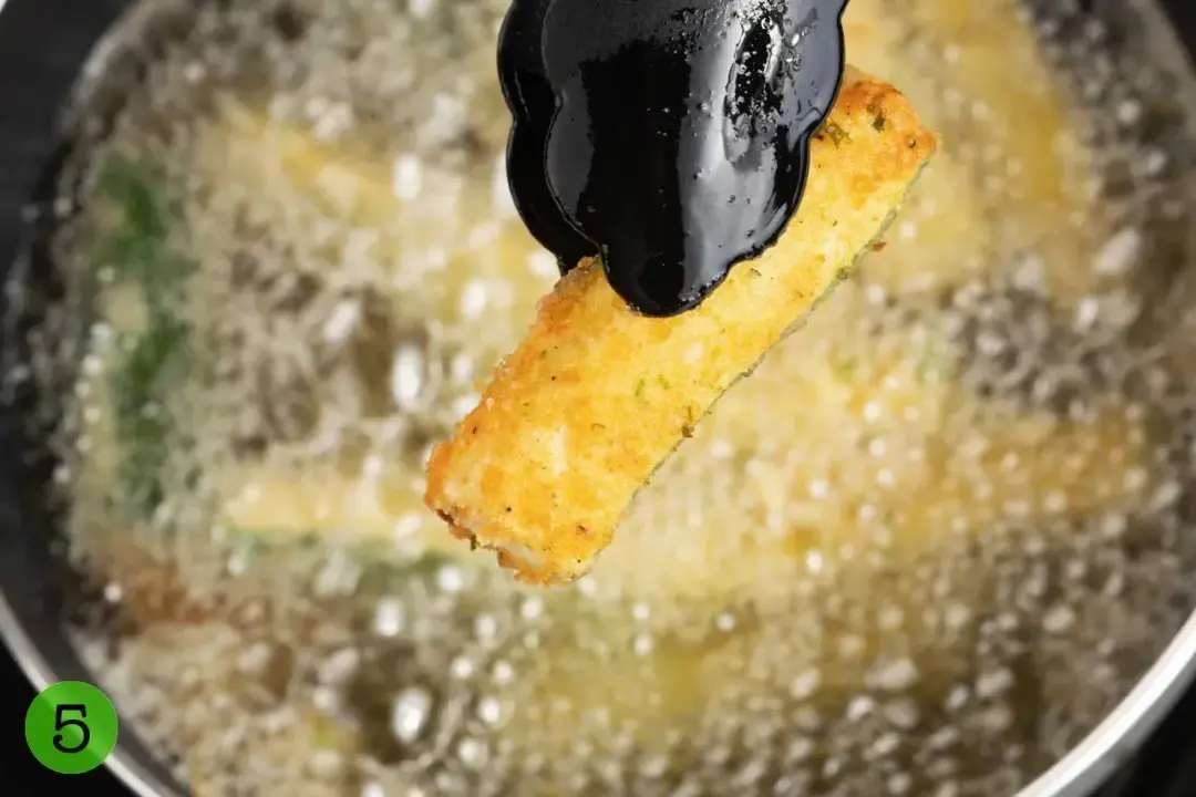 A fried breaded zucchini finger held above a sizzling pan of deep-frying zucchini fries