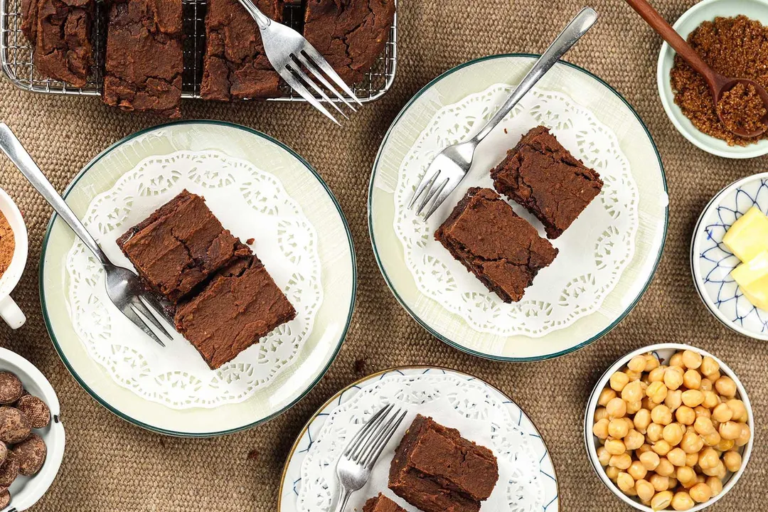 Plates holding chickpea brownie squares with forks on the side laid near a small disk of chickpeas, cocoa powder, and butter.