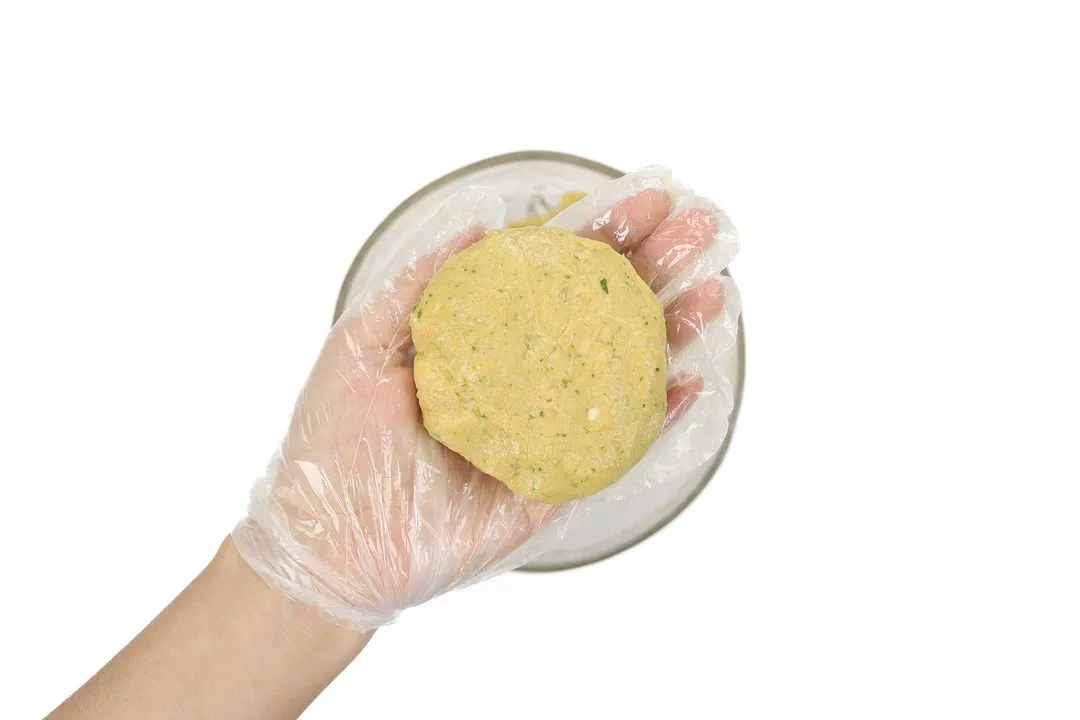 A hand wearing a disposable plastic glove holding a pattie of blended chickpeas.