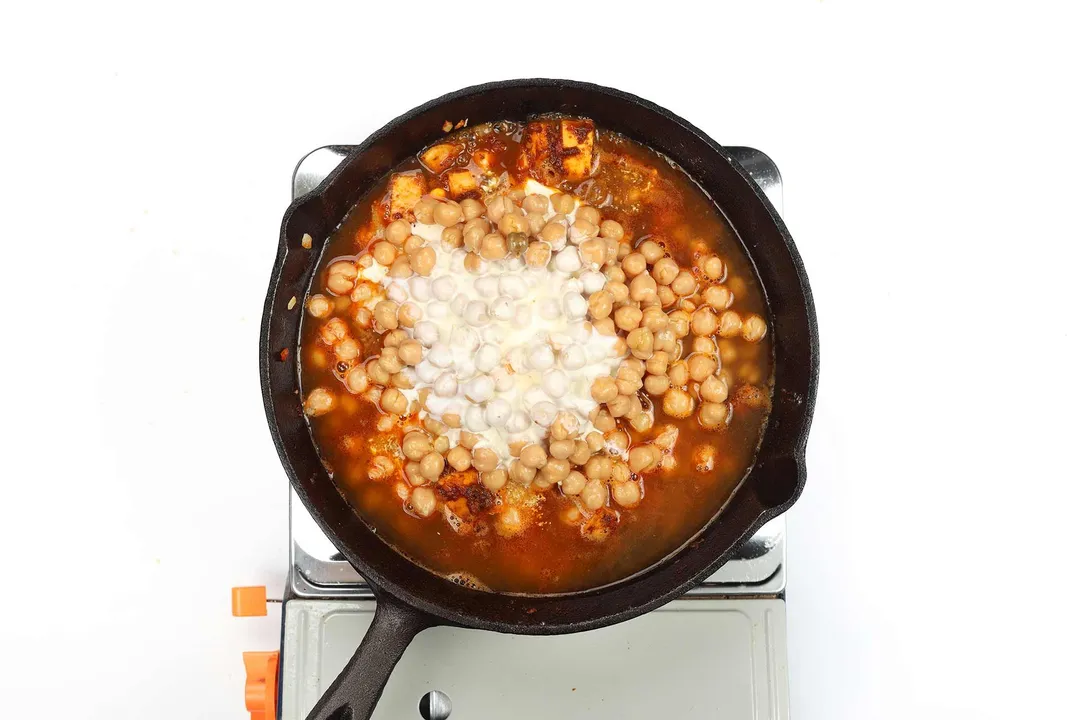 A skillet cooking chickpeas in a liquid mixture of curry and heavy cream on a portable gas stove.