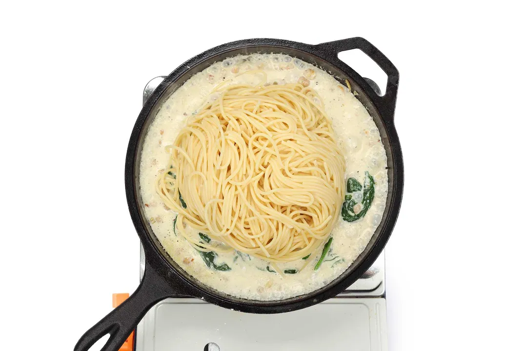 A skillet cooking spaghetti pasta in a cream sauce with some spinach on a portable gas stove.