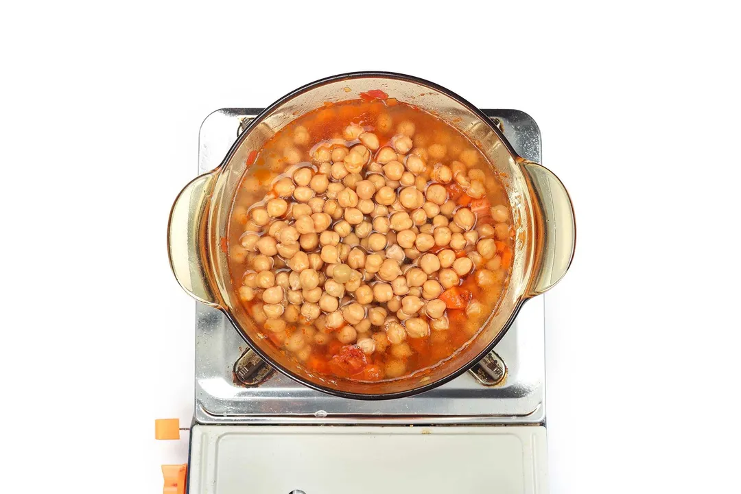 A saucepan cooking chickpeas in a broth on a portable gas stove.