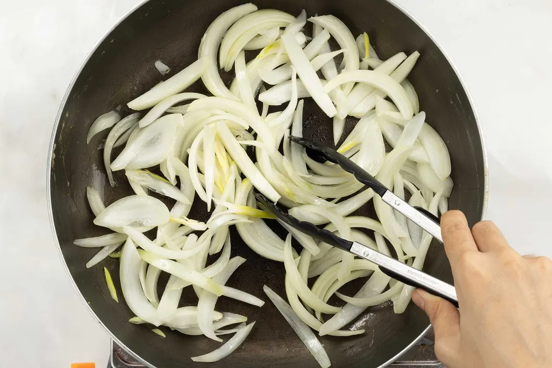 A hand stir-frying slices of onion on a hot pan.