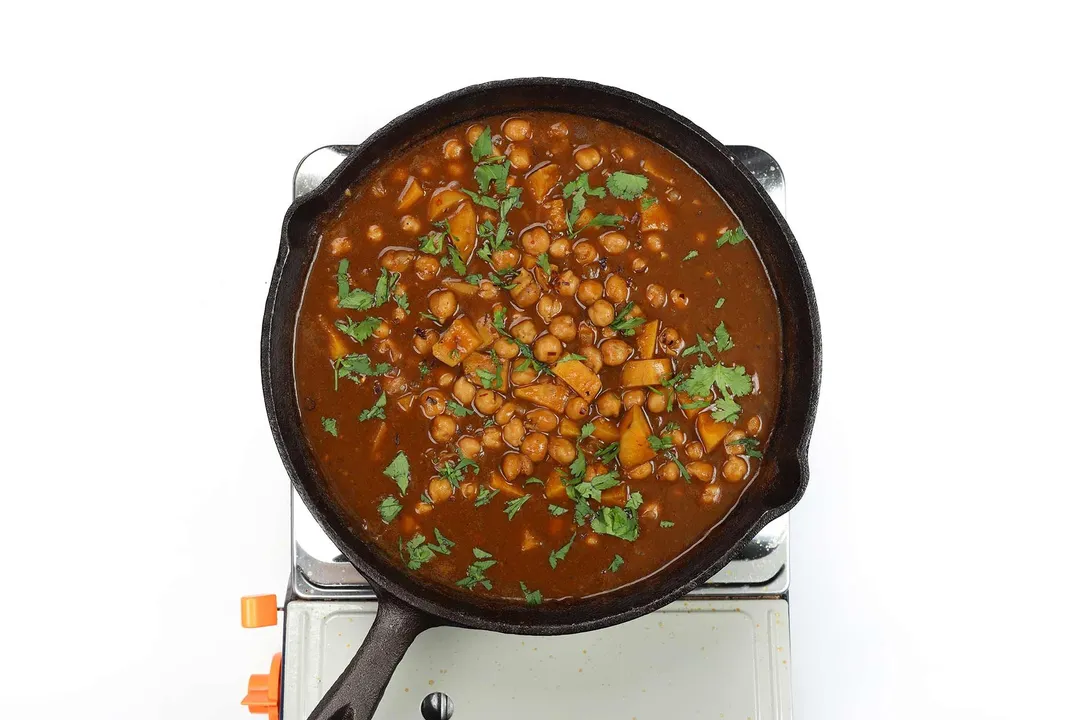 A skillet cooking chickpea curry with coriander leaves on a portable gas stove.