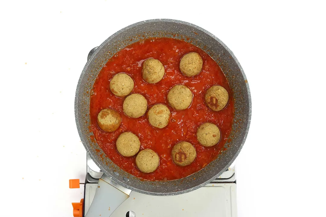 A pan cooking chickpea meatballs in tomato sauce on a portable gas stove.