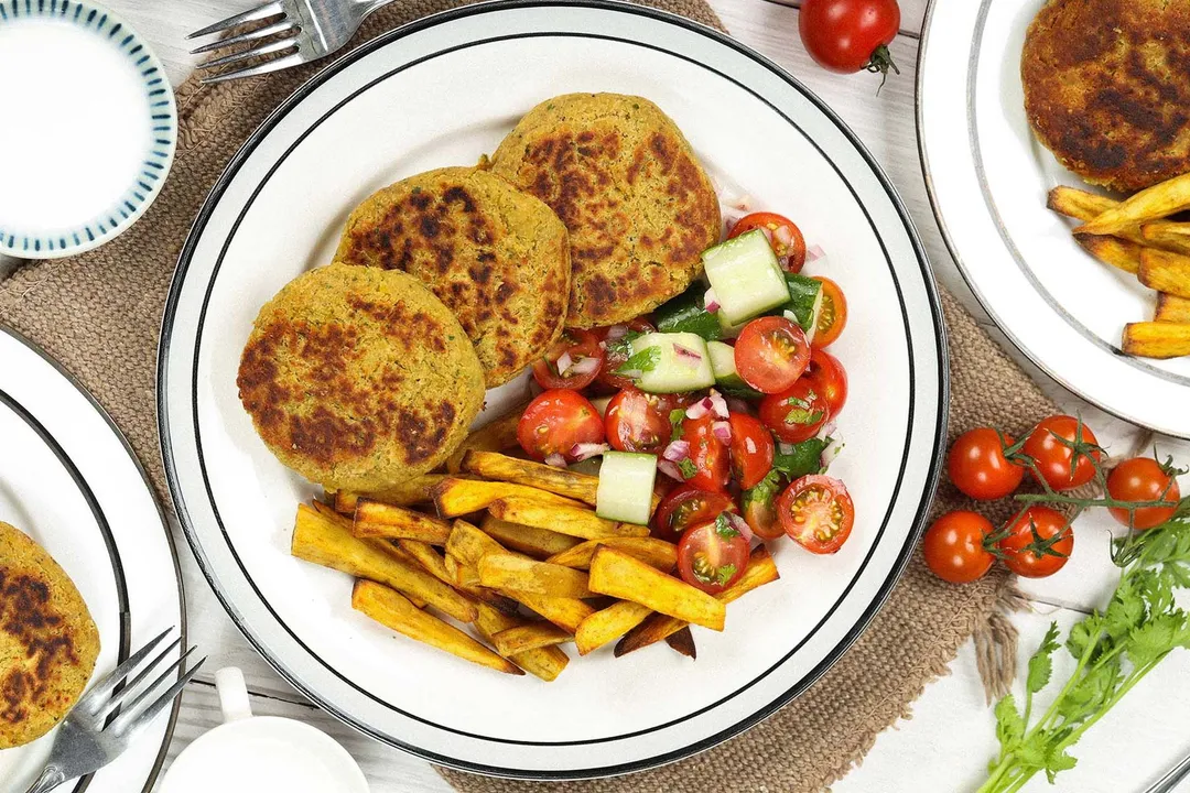 A round white plate filled with chickpea patties, air-fried sweet potato sticks, cherry tomato halves, and cucumber cubes.