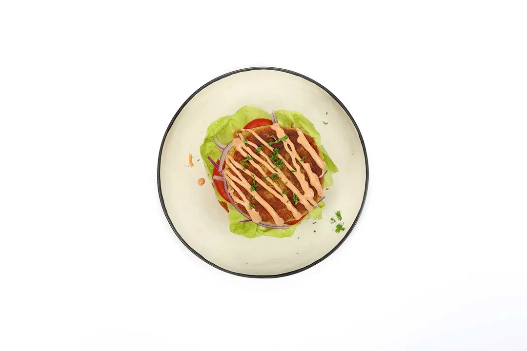 A chickpea pattie drizzled with a sauce stacked on top of lettuces and tomatoes, all laid on a round white plate.