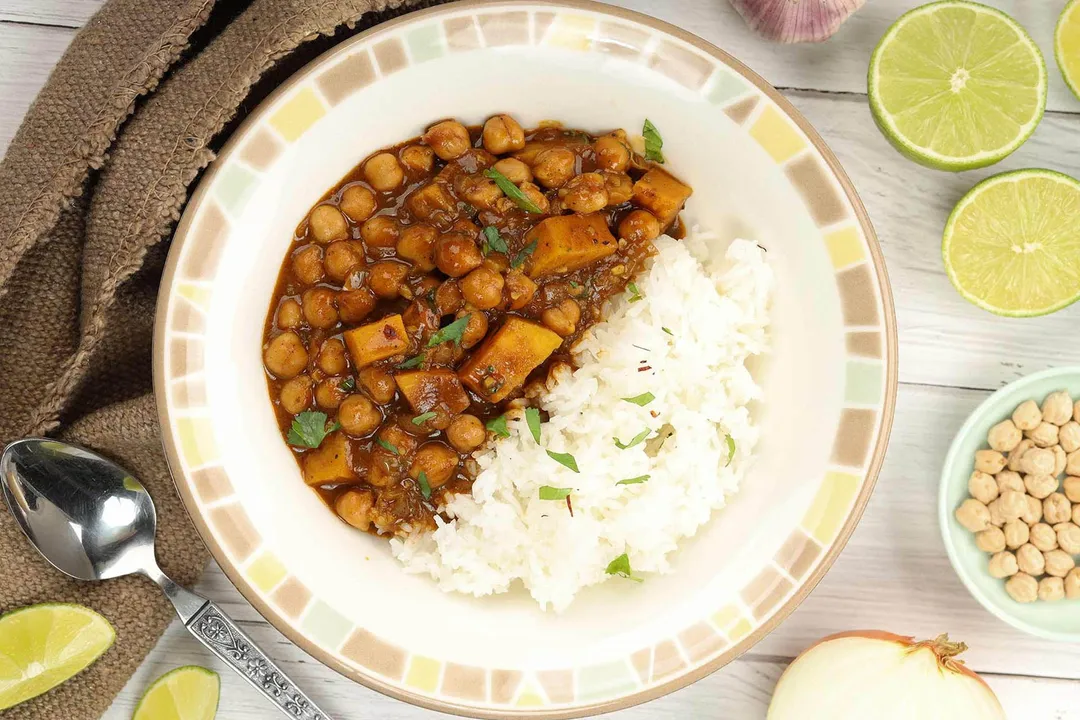 A plate of chickpea curry and white rice laid near a brown cloth, lime wedges, and a spoon.