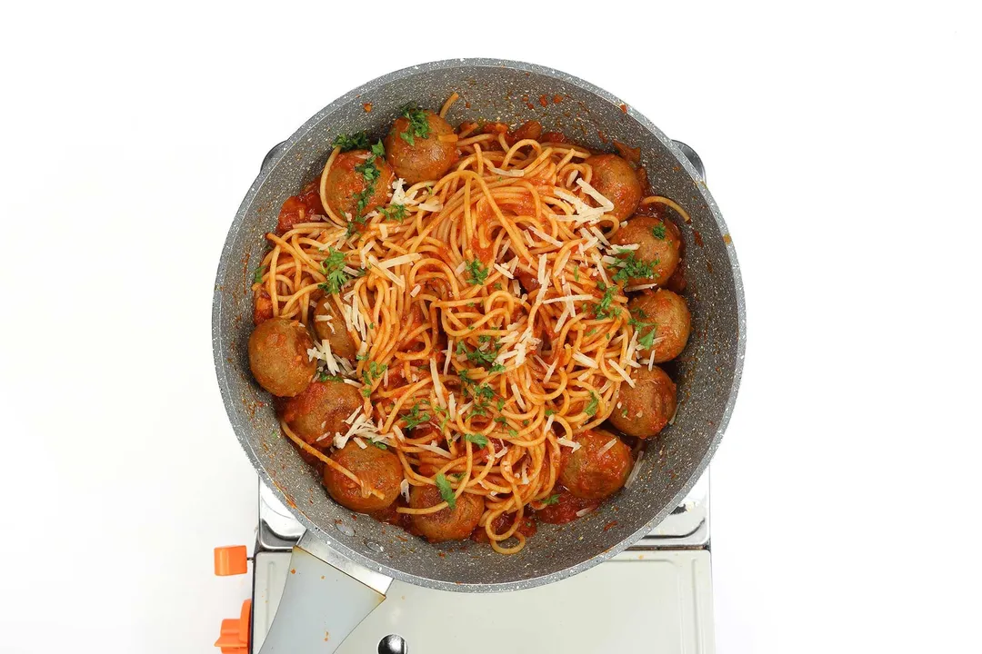 A pan cooking chickpea meatballs in tomato sauce and garnished with chopped parsley and shredded cheese on a portable gas stove.