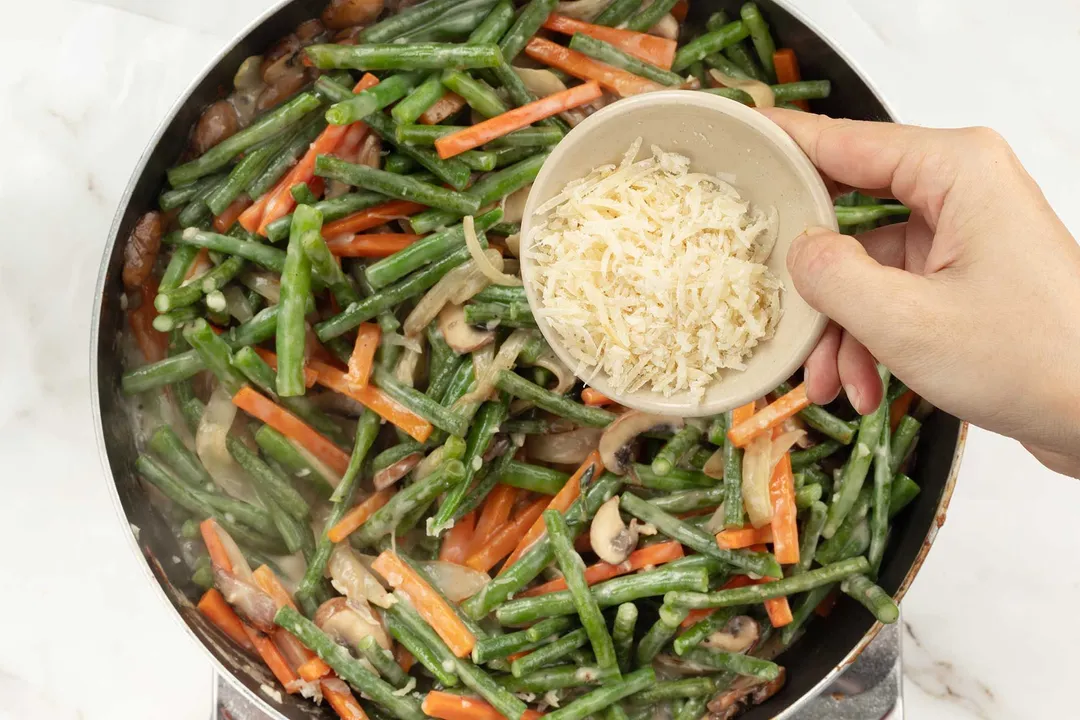 A hand adding shredded cheese into a pan filled with green beans and carrot matchsticks.