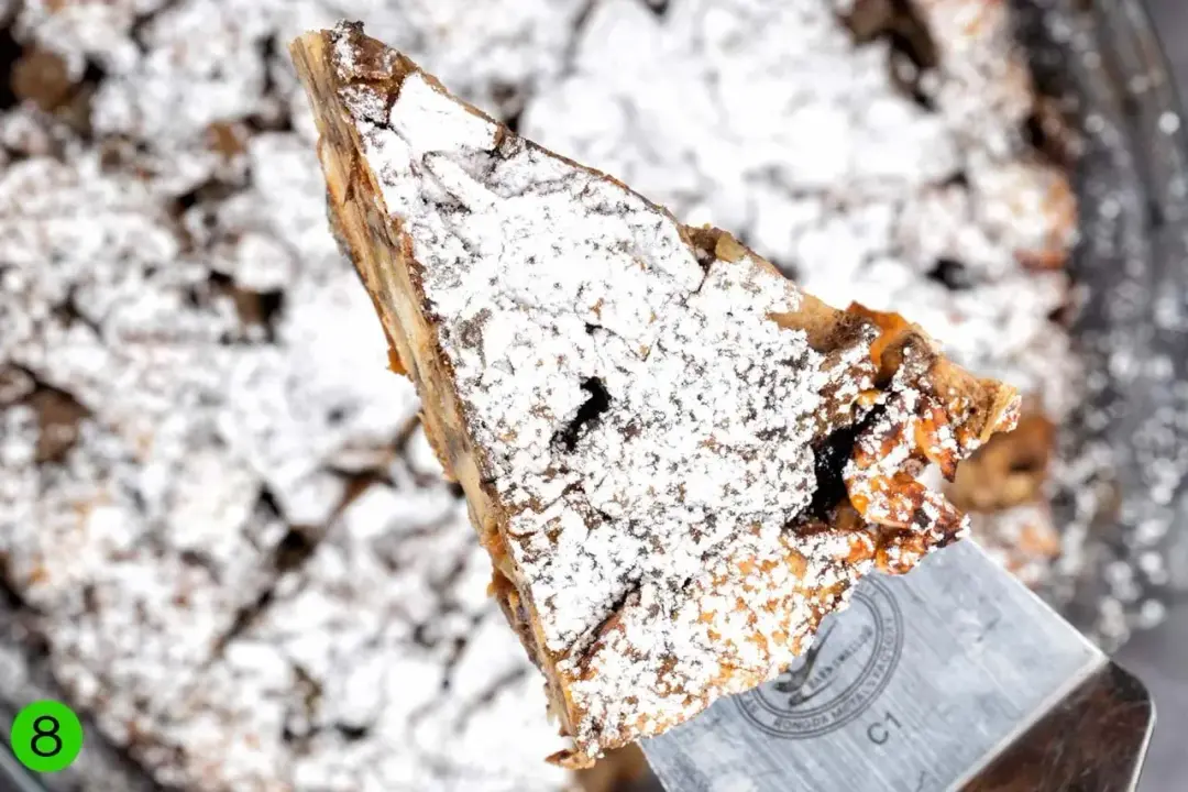 A slice of French toast casserole hovered over a round baking dish, fully dusted with powdered sugar