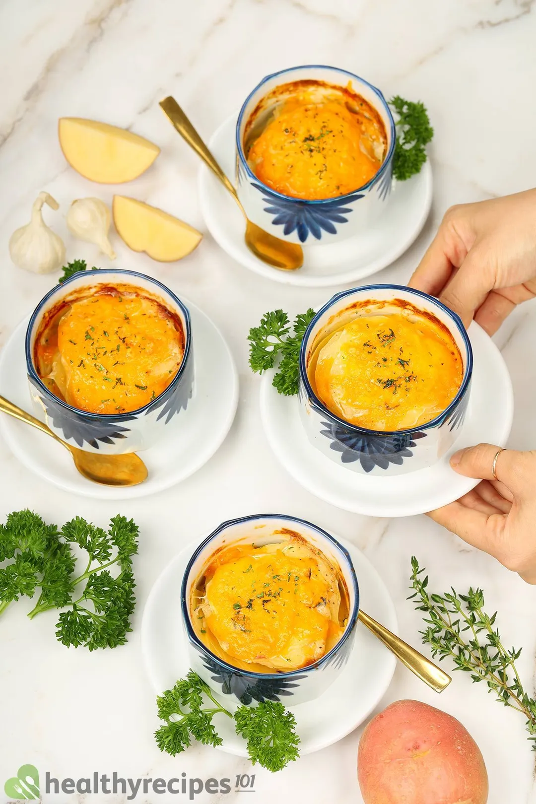 Four bowls of potato gratin laid near fresh parsley and potatoes, one of which is being picked up by a pair of hands.