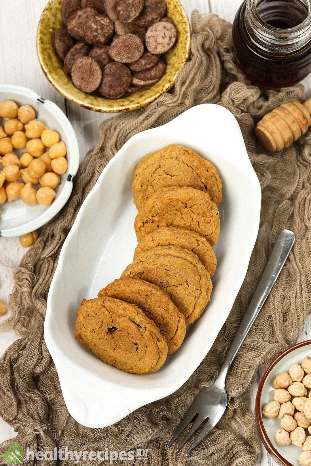 A small, rectangle baking dish holding chickpea cookies laid near a small bowl of chocolate chips, a bowl of chickpeas, and a jar of honey.