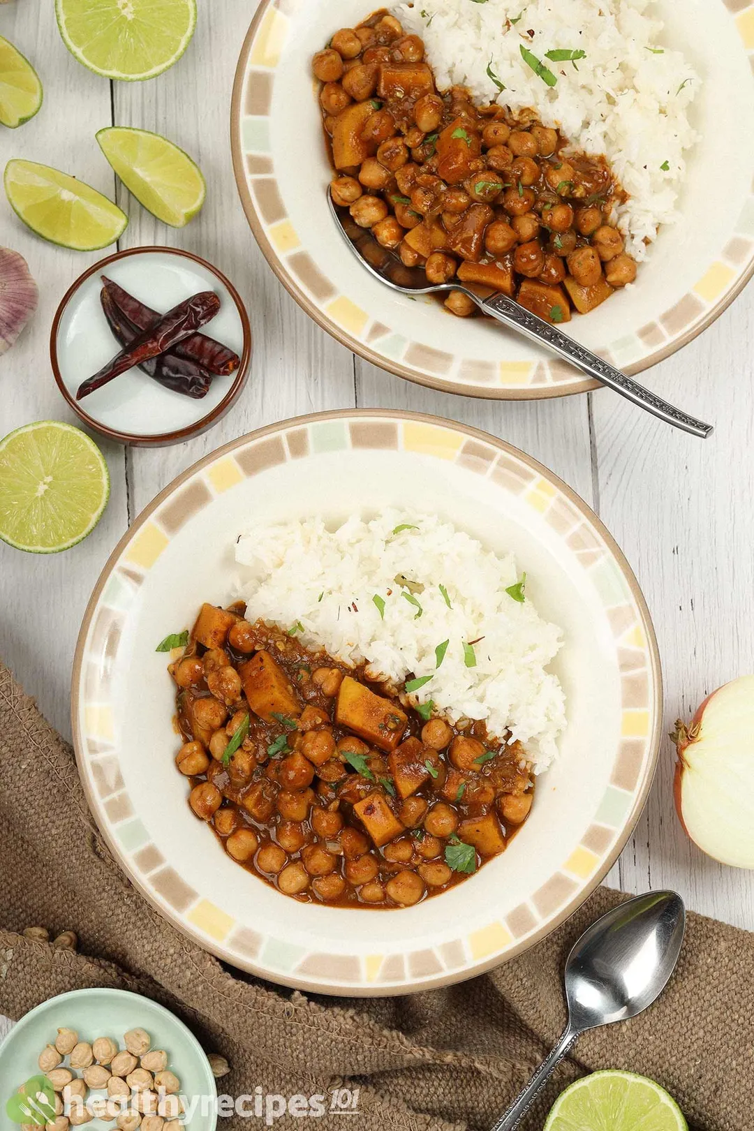 Two plates of chickpea curry laid near a brown cloth, spoons, lime wedges, and dried chili.