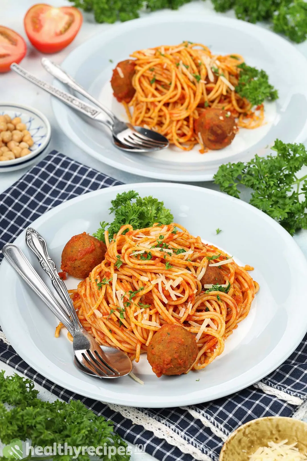 Two plates of chickpea meatball spaghetti with spoons and forks on the side, laid near parsley leaves.
