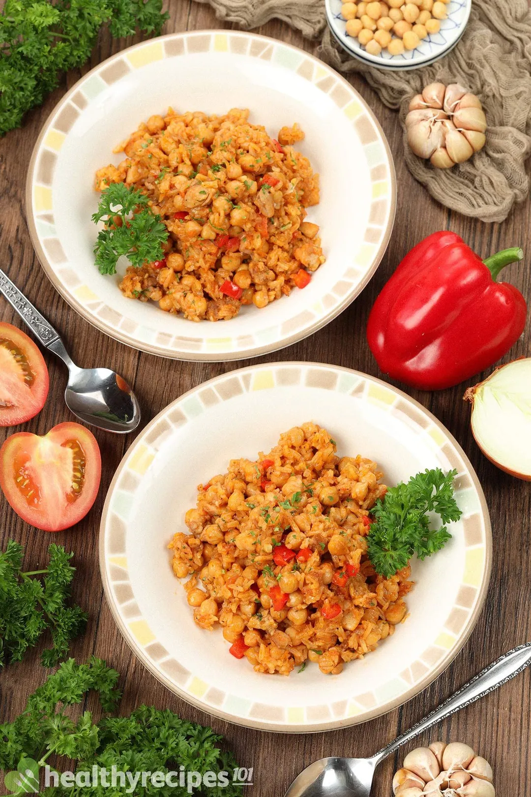 Two plates of chickpea rice surrounded by a red bell pepper, tomato halves, fresh parsley, garlic cloves, and spoons.