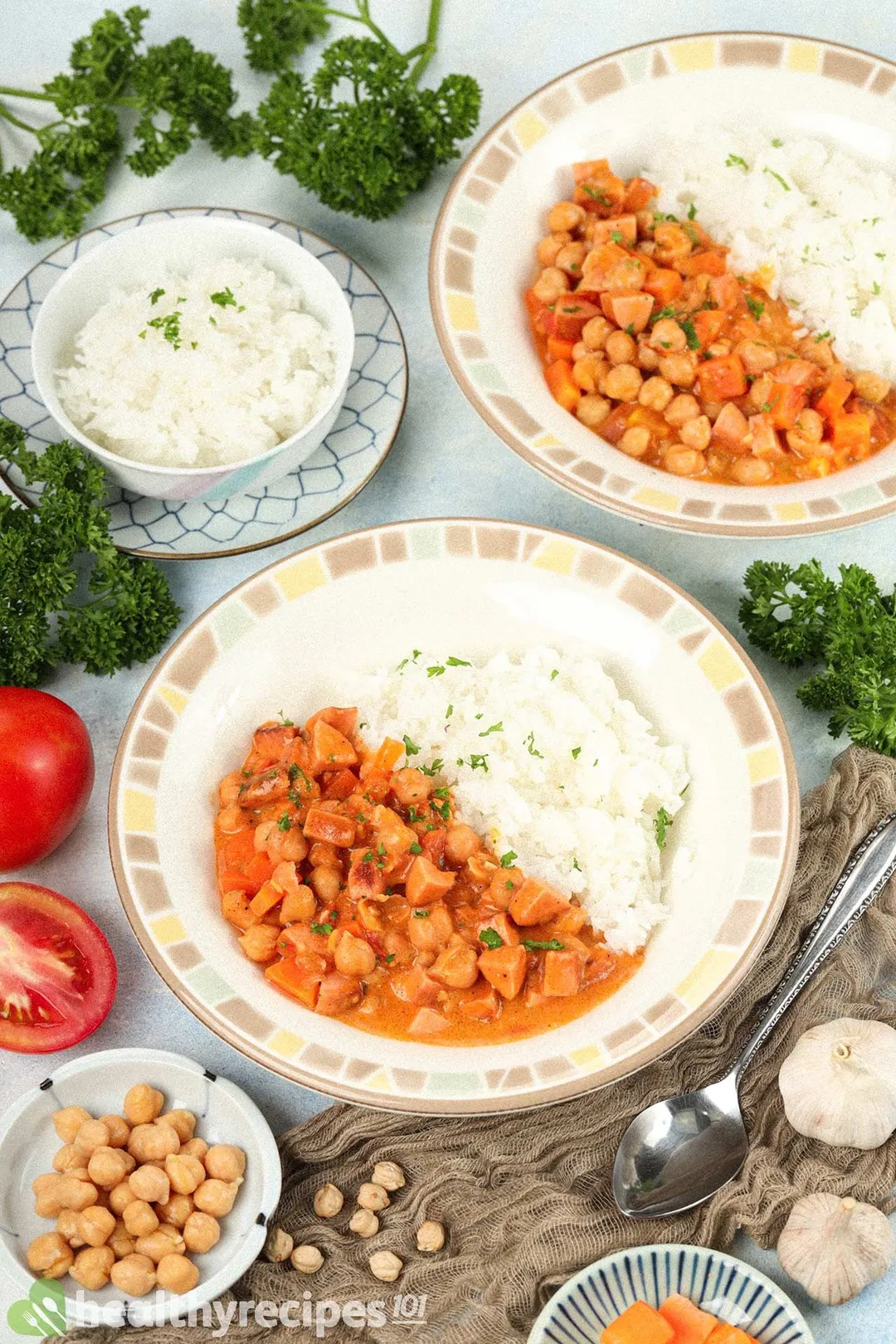 Plates of chickpea stew and cooked white rice surrounded by a small disk of chickpeas, a small bowl of cooked white rice, fresh parsley, and tomatoes.