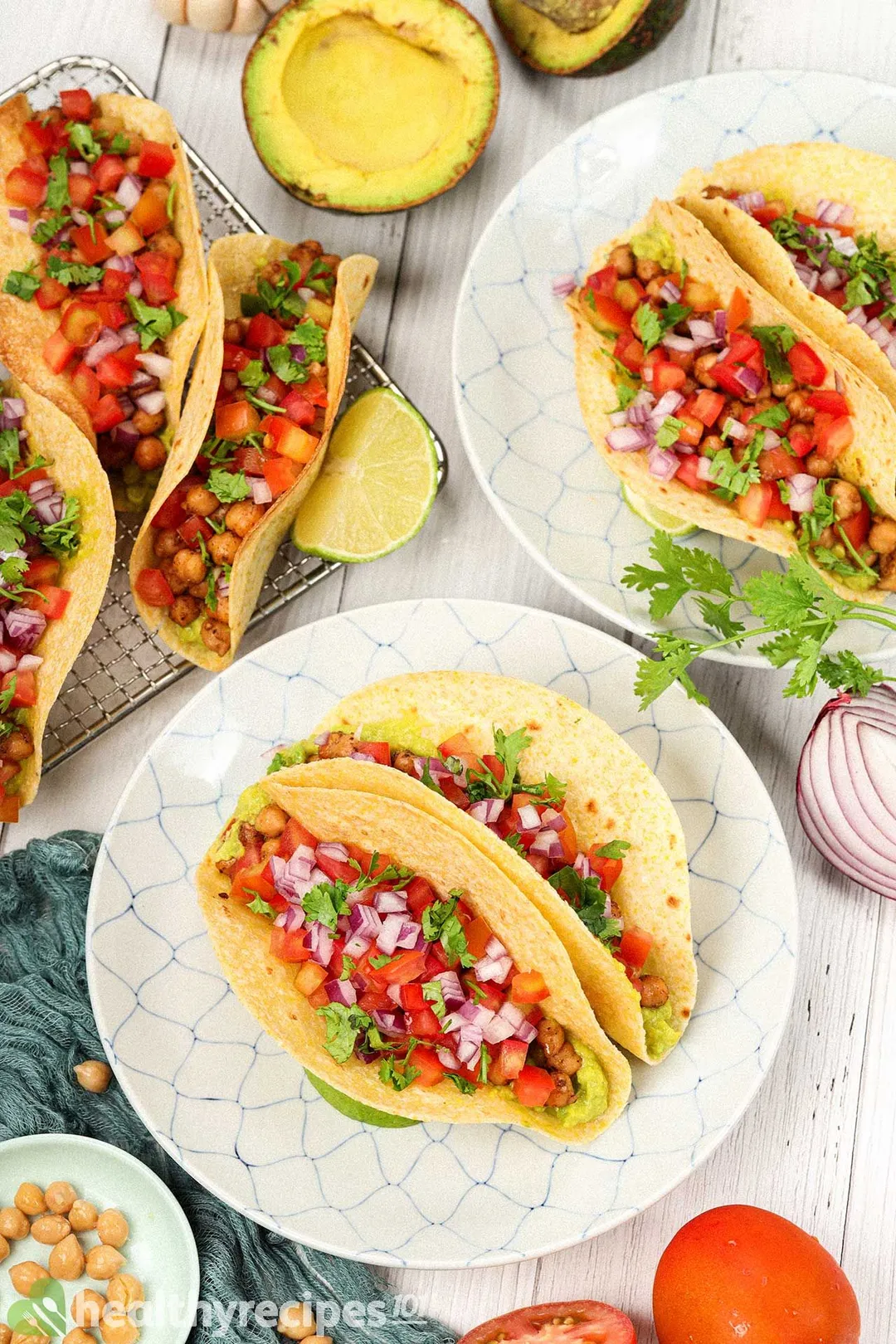 Plates of chickpea tacos laid near a tomato, coriander, avocado halves, and a small disk of chickpeas.