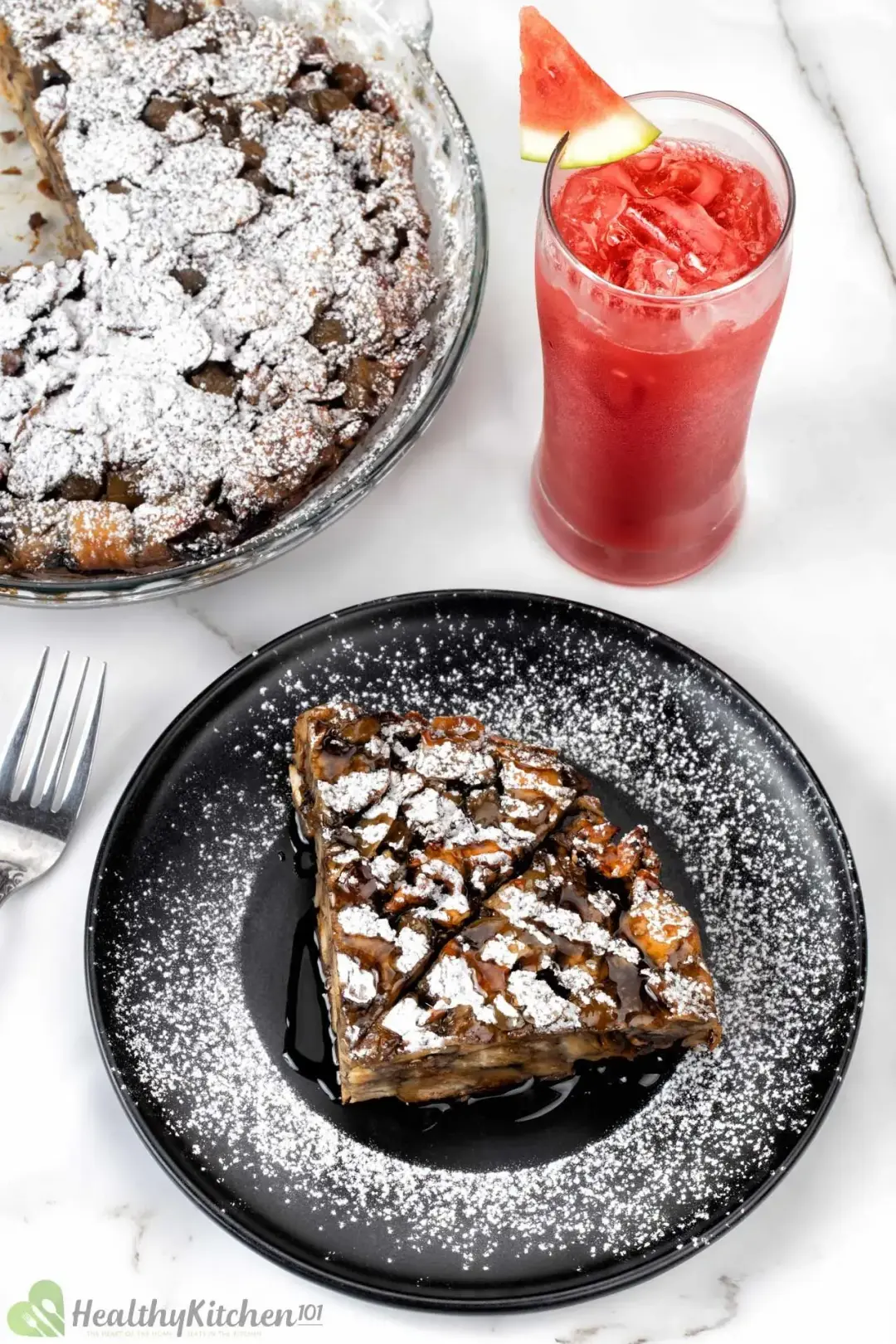 A black plate of French toast casserole dusted with powdered sugar, next to a large sweet casserole and iced watermelon juice