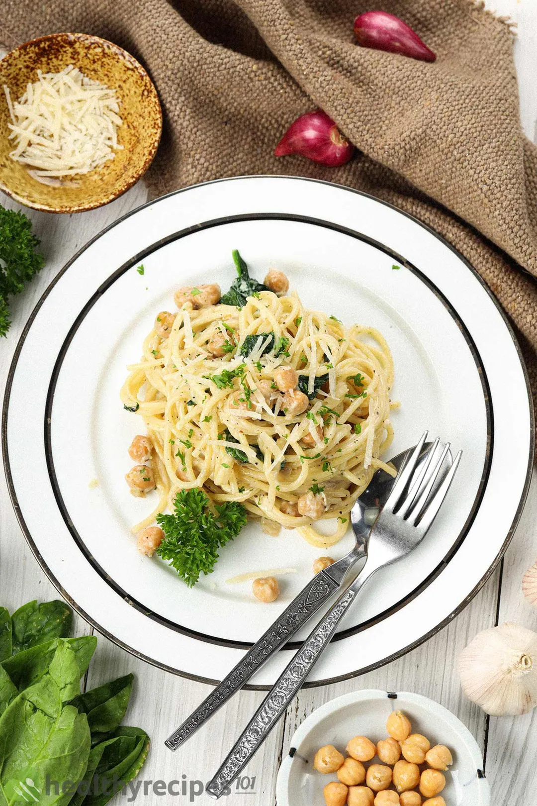 A round white plate containing chickpea pasta, a fork, and a spoon laid near a brown cloth, shallots, and garlic cloves