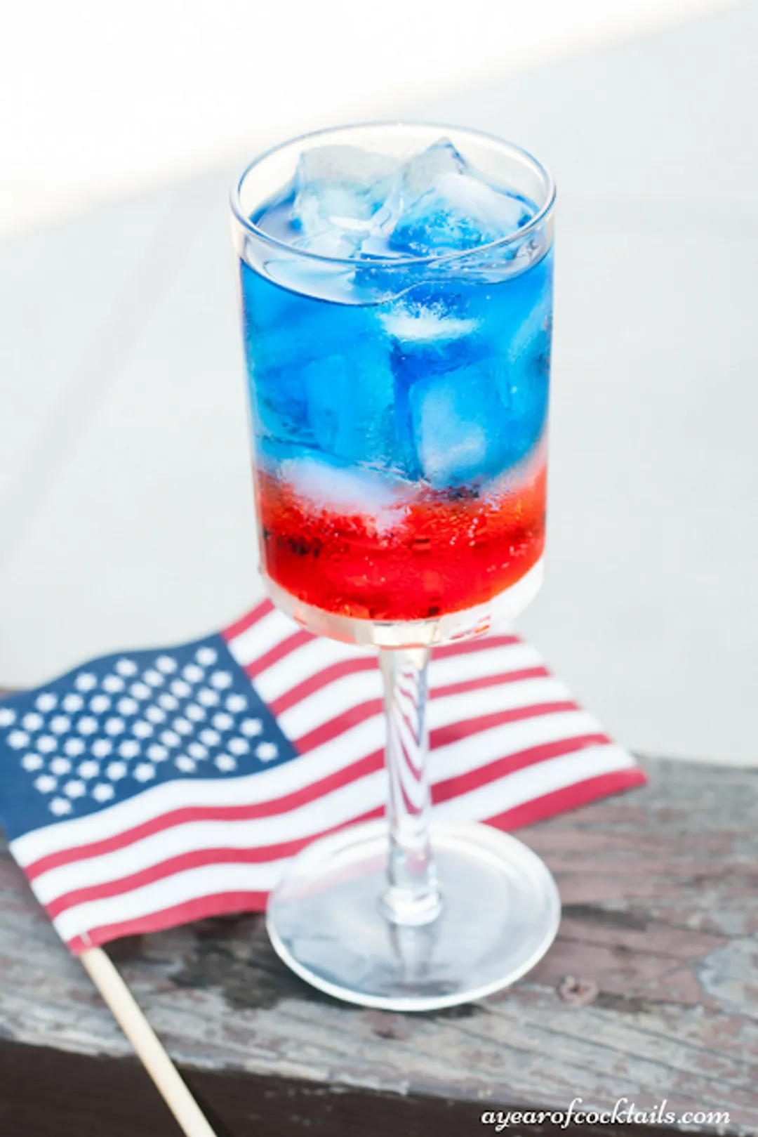 A glass filled with a bright red drink, garnished with a slice of lime and a sprig of rosemary. The flag of united state in the background