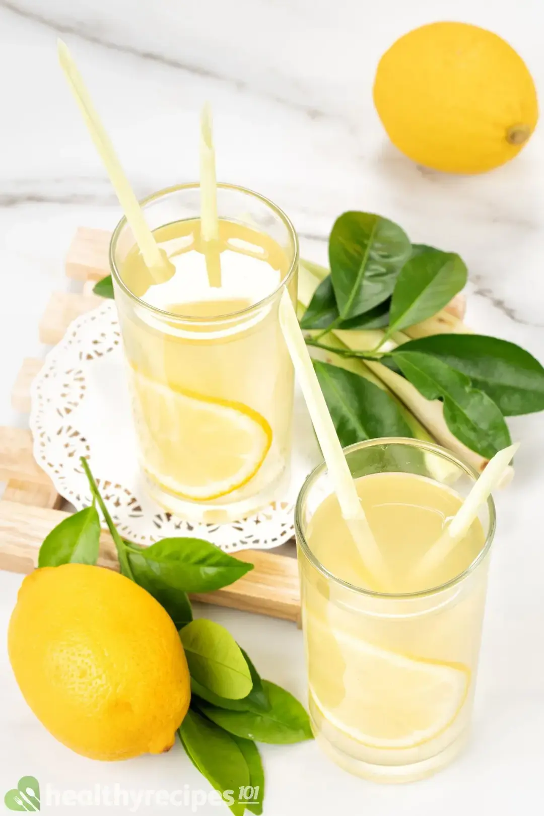 Two glasses of vinegar and lemon juice drinks, with white straws, whole lemons, and lemon leaves around