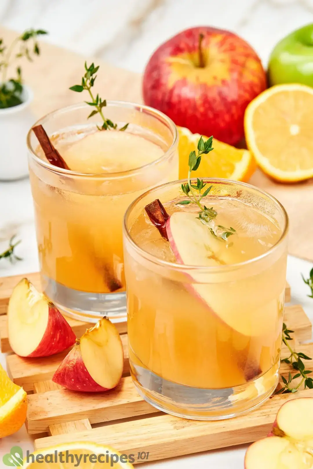 Two short glasses of apple juice cocktail with apple wedges, oranges, cinnamon, and rosemary sprigs