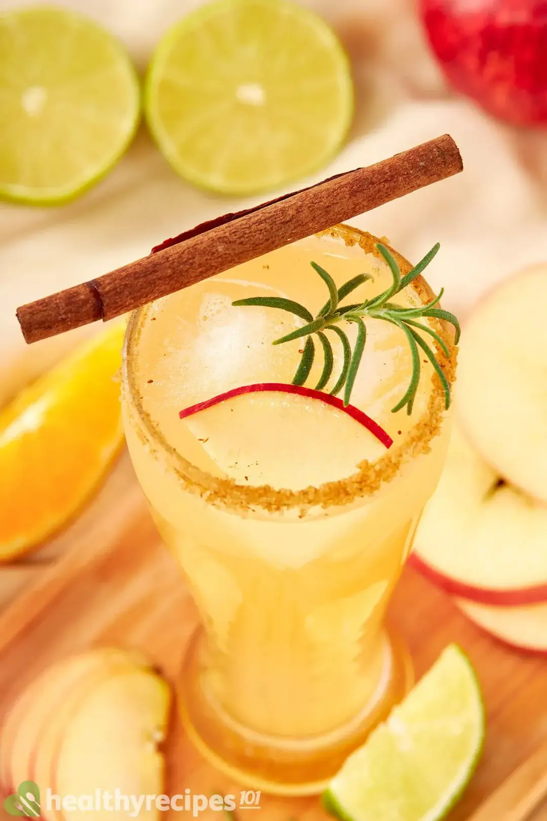 A tall glass of apple cider cocktail garnished on top with a cinnamon stick, a rosemary sprig, and apple wedges all around
