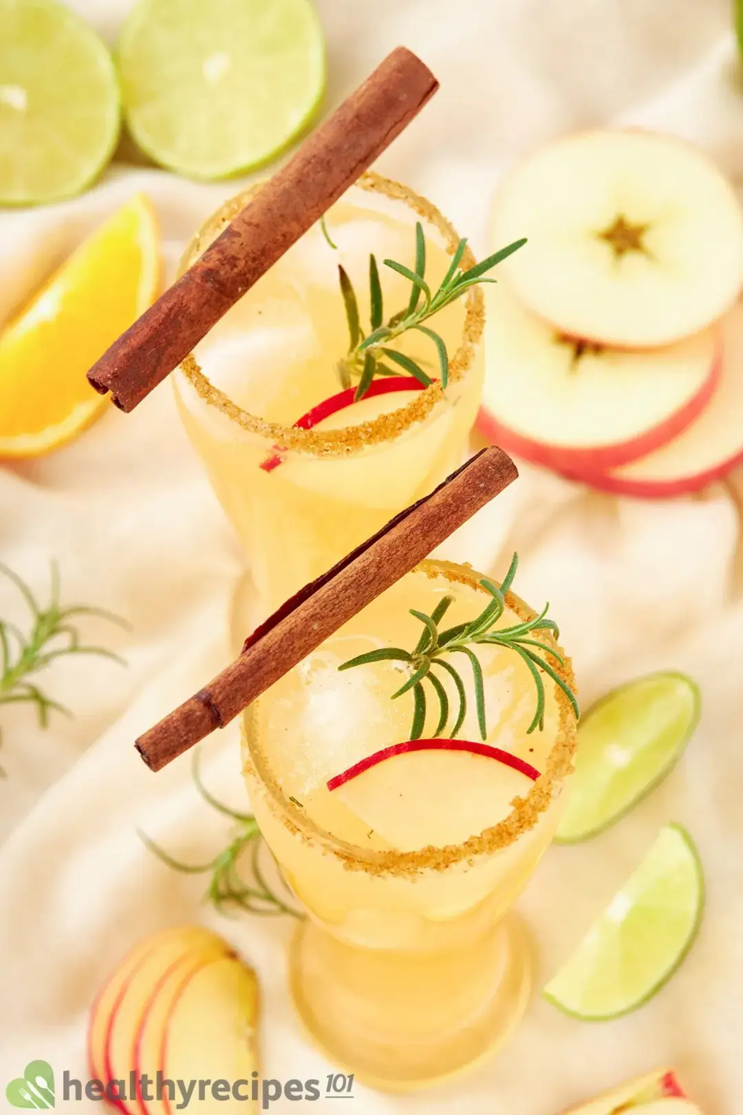 Two glasses of apple cider margarita cocktail garnished with cinnamon sticks, apple slices, lem on wedges, and rosemary sprigs