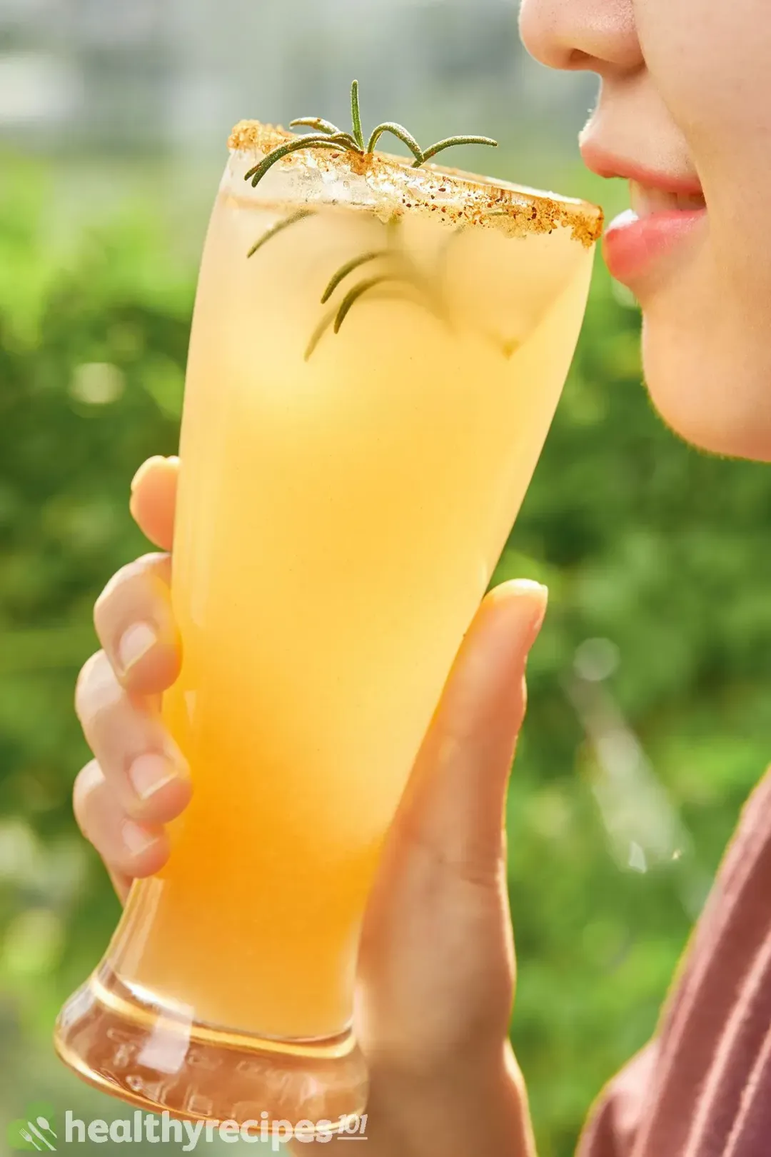 A person holding a tall glass of apple cider margarita with rosemary to drink