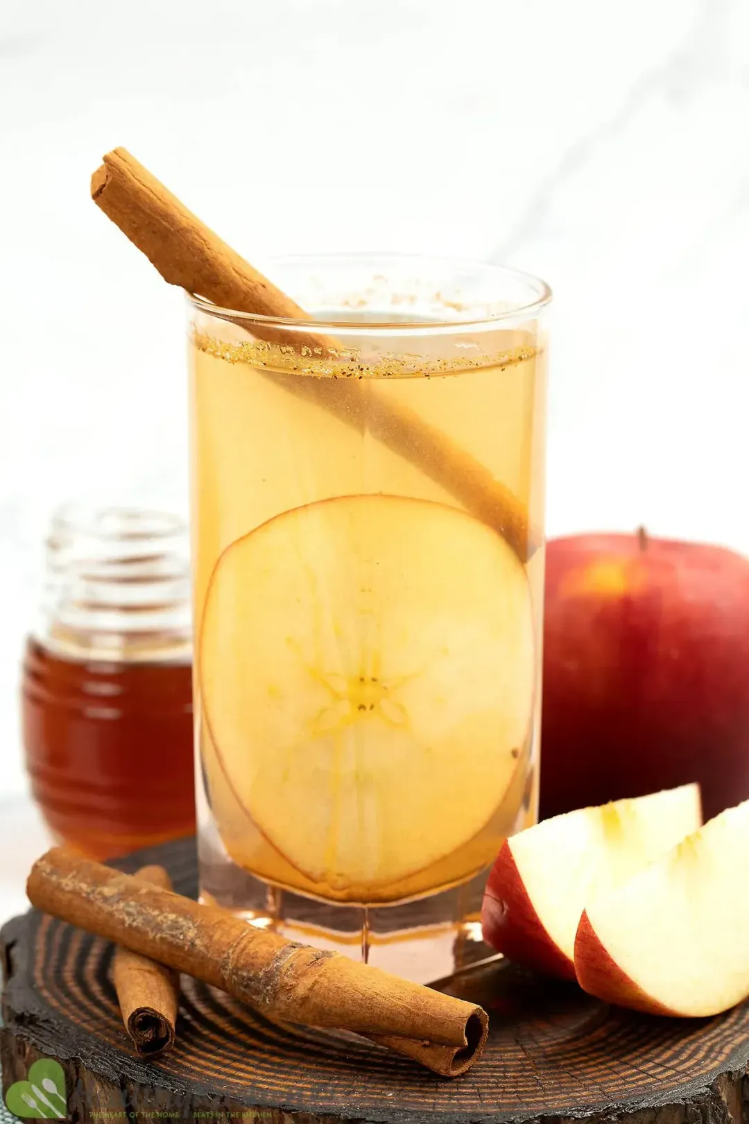 A shot glass of apple vinegar honey with cinnamon sticks, red apple wedges, and a honey jar