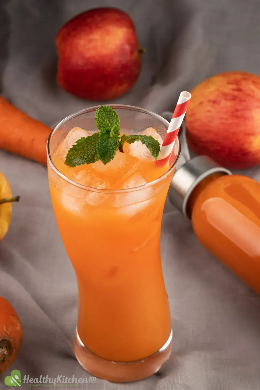 A glass of carrot juice with ice and mints, surrounded by red apples and a bottle of carrot juice