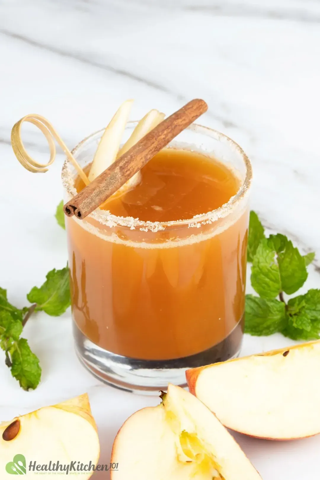 A glass of apple juice and vodka cocktail, topped with a cinnamon stick, next to mints and apple wedges