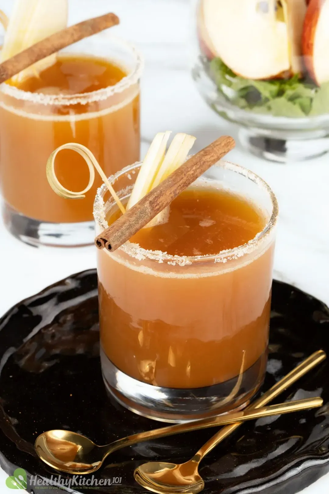 Two glasses of apple juice and vodka cocktail, garnished with cinnamon sticks and sliced apples, put on a black tray with gold spoons