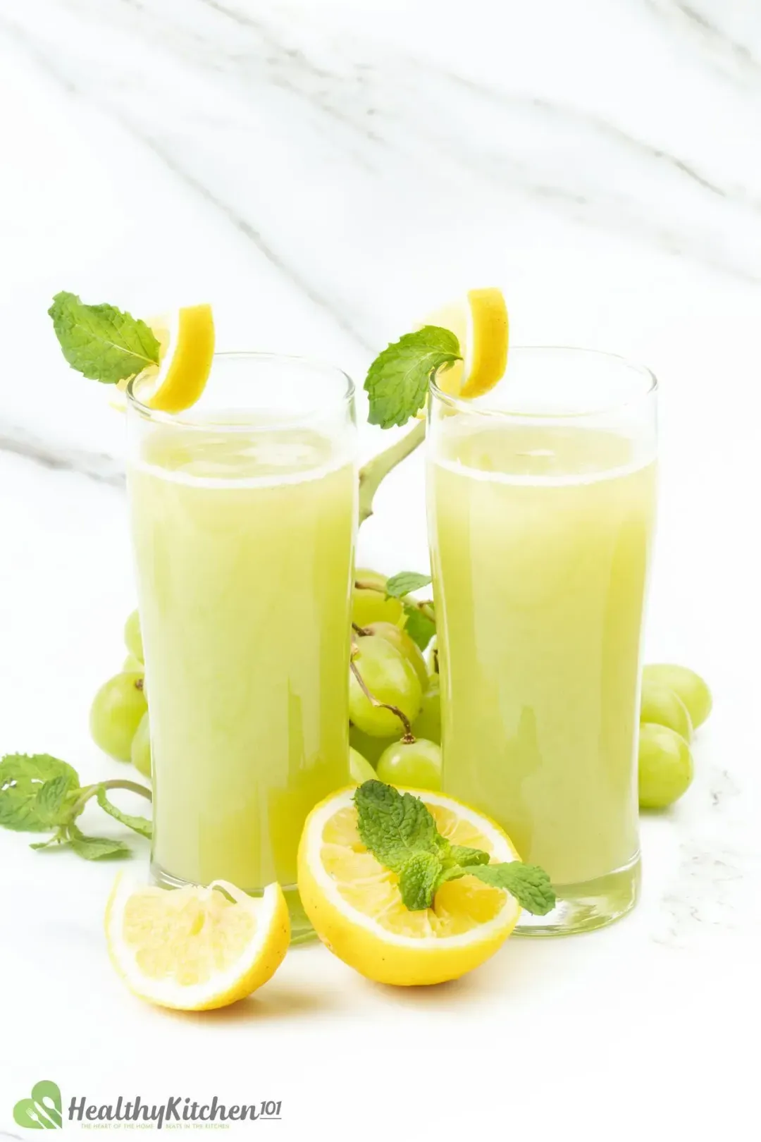 Two glasses of green grape juice placed side by side surrounded by some lemons and green grapes