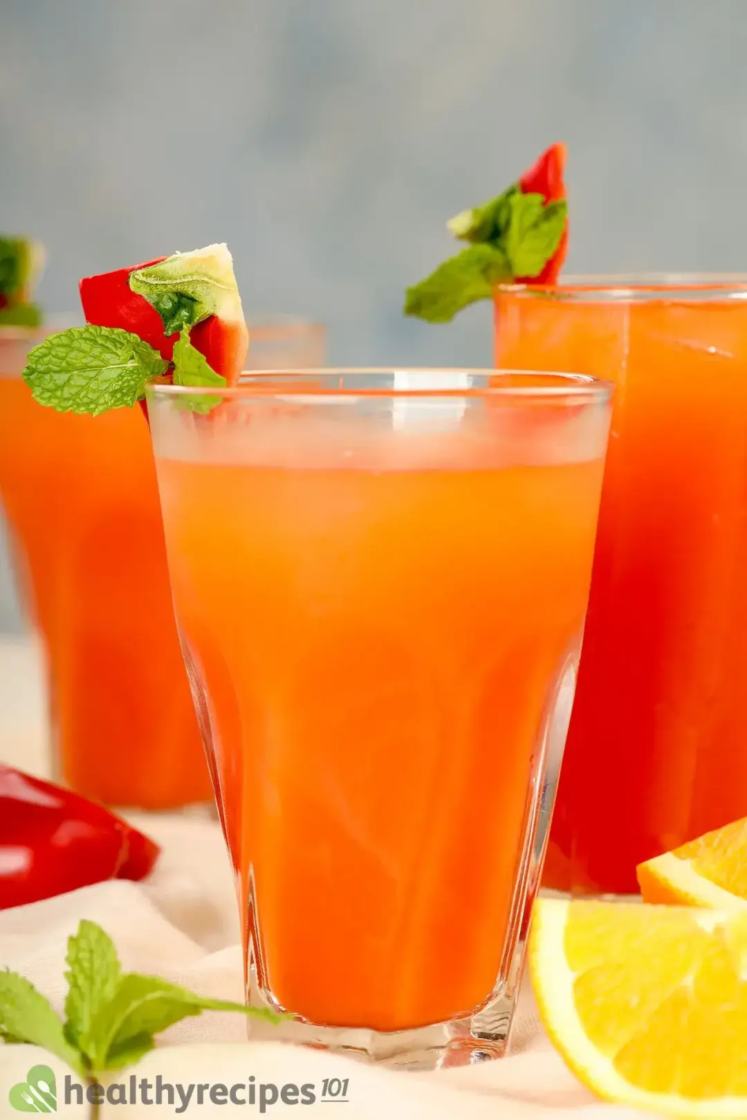 Three glasses of Bell Pepper Juice on a table decorated with orange sliced, half bell pepper, mint leaf
