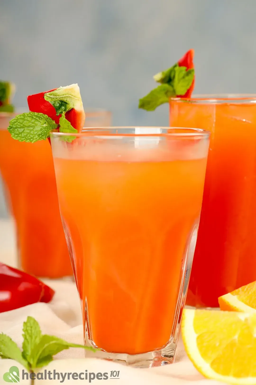 Three glasses of Bell Pepper Juice on a table decorated with orange sliced, half bell pepper, mint leaf