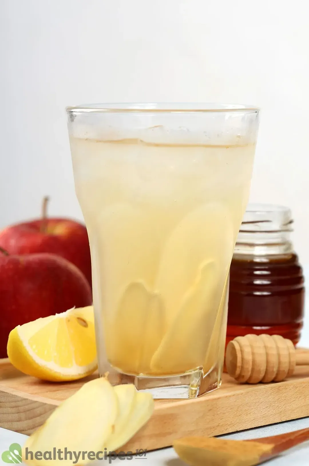 A large glass of apple cider vinegar drink filled with ginger slices, next to ginger slices and a jar of honey