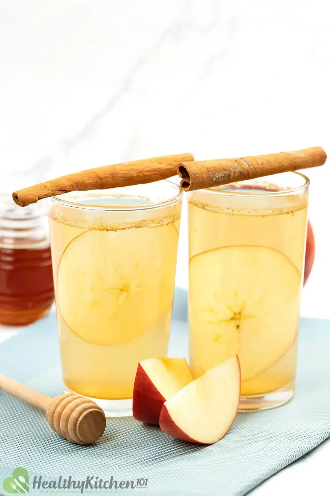 two glasses of apple cider vinegar and honey side by side with cinnamon sticks on top, decorated with apple sliced, and honey jar